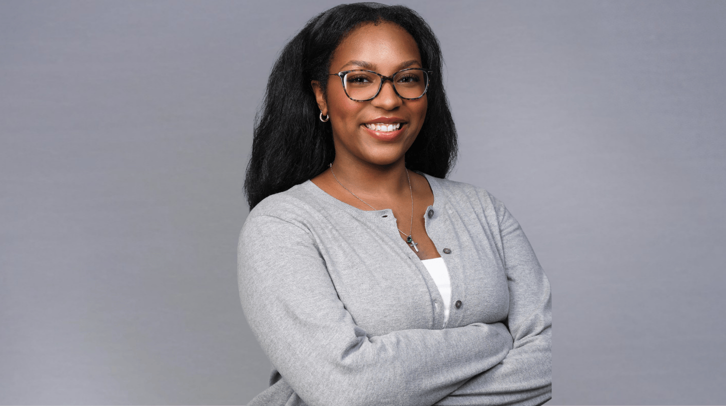 Portrait of a current Gannon Scholar with long dark hair and glasses, smiling, wearing a gray cardigan over a white top and a cross necklace, standing against a neutral gray background with arms crossed.